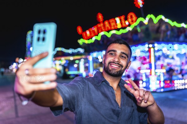 Happy young man smiling and taking a selfie while gesturing a peace sign, surrounded by vibrant lights at an amusement park at night