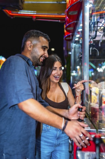 Young couple having fun playing a claw crane game at an amusement park at night, trying to win a prize