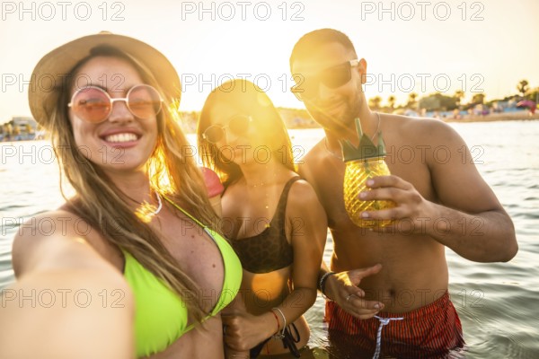 Three cheerful friends are taking a selfie while having a refreshing cocktail in the sea during a summer sunset
