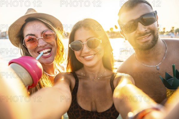 Group of cheerful multi ethnic friends taking selfie at beach party, enjoying refreshing drinks during sunset