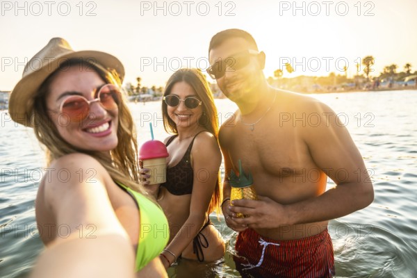 Group of cheerful young friends taking selfie in the sea while drinking cocktails at sunset during summer vacation