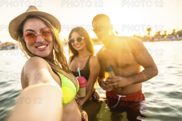 Three friends are taking a selfie while enjoying drinks in the sea during a beach party at sunset