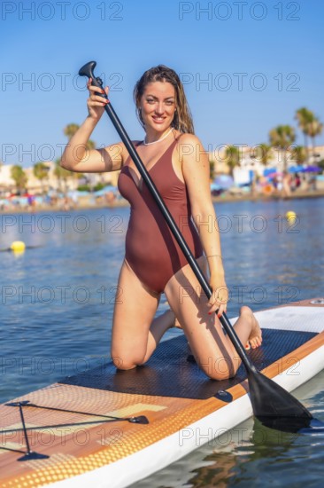 Woman kneeling on a paddleboard, enjoying a sunny day at the beach. Clear blue sky and palm trees in the background enhance the summer vibe