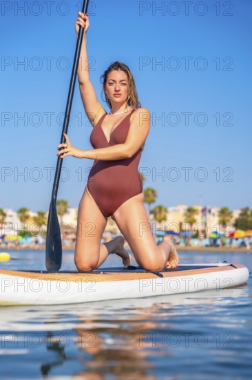 Young woman in a swimsuit kneels on a paddle board, holding a paddle and enjoying a summer day on the water