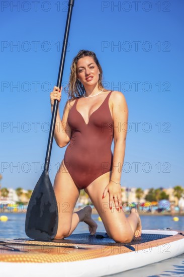 Young woman kneeling on a paddle board, holding a paddle and enjoying a summer day at the beach
