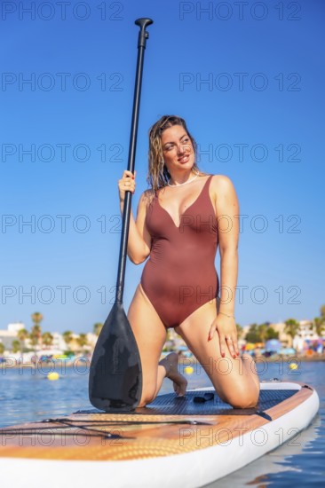 Young woman kneeling on a paddle board, holding a paddle and enjoying a summer vacation at the beach