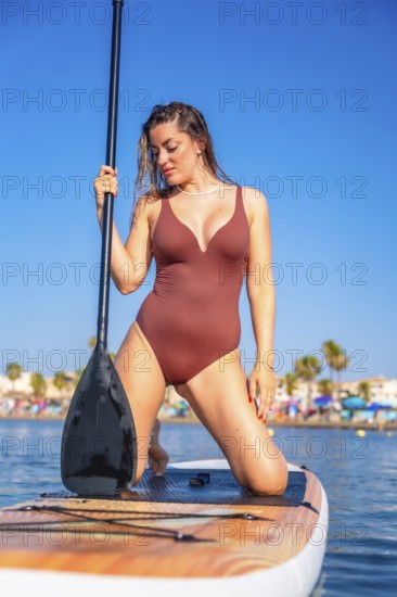 Young woman kneeling on a paddle board, holding a paddle, enjoying a sunny summer day at the beach