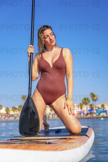 Young woman kneeling on paddle board, holding paddle and looking away, enjoying summer sunny day at the beach