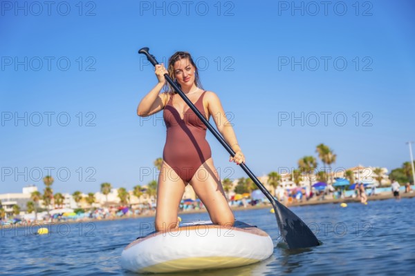 Young woman kneeling on a paddle board, paddling in the sea near the beach on a sunny summer day