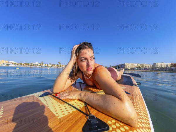 Young woman lying on a paddle board, soaking up the sun and enjoying the serene beauty of the calm sea along the coast