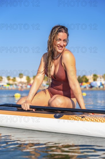 Woman enjoying paddle surfing on a calm sea, smiling under the bright summer sun, with a vibrant beach background full of activity