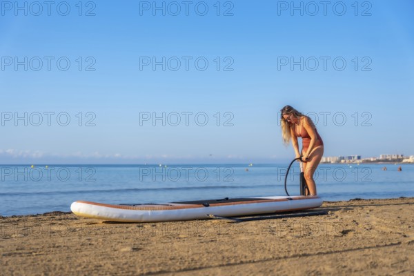 Woman inflating a paddleboard on a sandy beach, ready for a summer day of paddle surfing under a clear blue sky