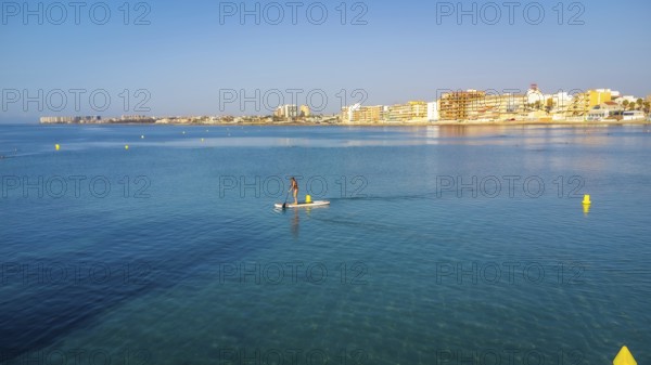 Calm waters and clear skies set the scene for a serene paddle boarding experience near a coastal town, highlighting summer leisure and tranquility