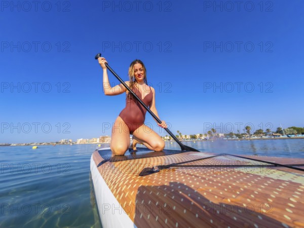 Young woman kneeling on a paddle board, holding a paddle, enjoying a summer watersport activity on calm blue water