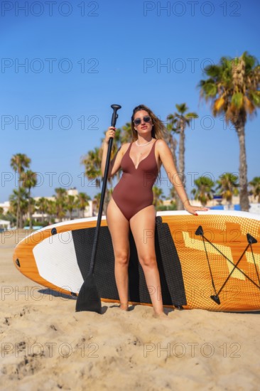 Young woman in swimsuit holding a paddle and posing with her paddle board on the beach on a sunny summer day