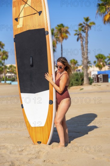 Young woman holding a paddle board on a sandy beach with palm trees in the background, enjoying summer vacation
