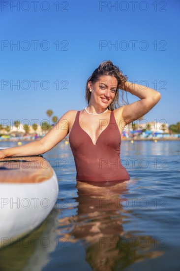 Young woman enjoying summer vacation, posing in the sea next to her paddle board, wearing a swimsuit