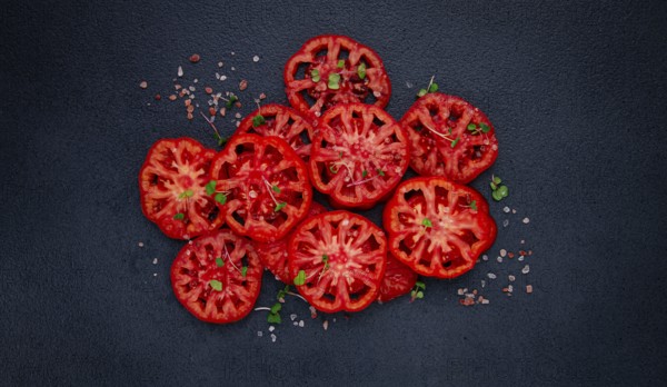 Freshly sliced ripe tomatoes arranged on a textured dark surface with herbs and salt scattered around