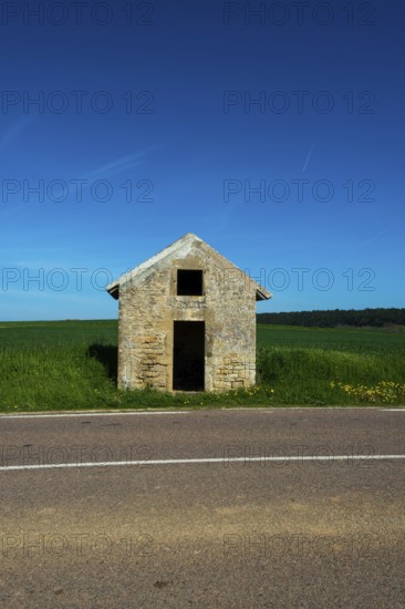 A rustic stone hut stands alone by a quiet road, surrounded by vibrant green fields. Auvergne. France