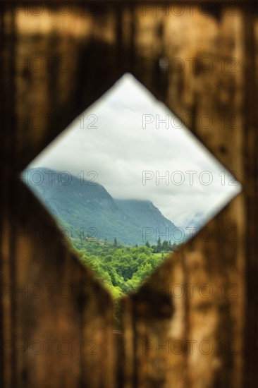 Rustic wooden frame reveals a lush green landscape with mountains in the background under a cloudy sky. France