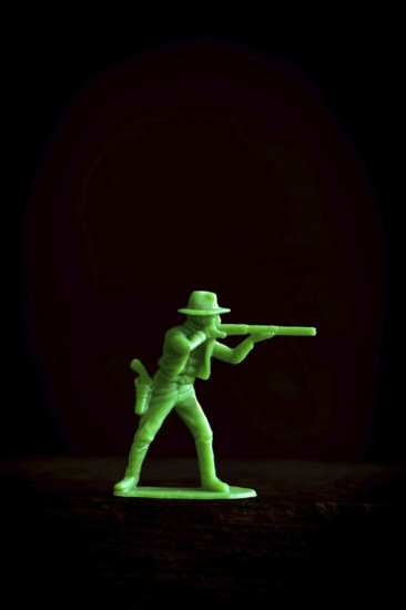A vibrant green plastic toy cowboy stands in a tactical pose, aiming a rifle against a dark background. The scene highlights the toy's details and engages with playful nostalgia