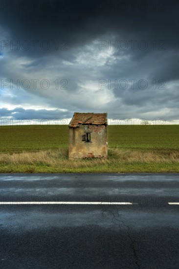 A small, dilapidated hut with a broken roof and cracked walls is positioned next to a quiet road. Dark clouds gather above, Puy de Dome, France