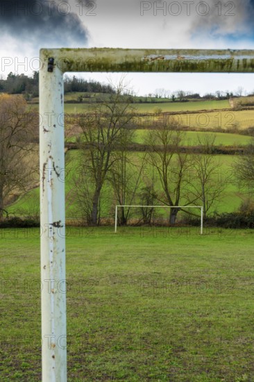 A weathered soccer goalpost stands in the foreground of a wide grassy field, with lush green hills and trees in the background, Auvergne. France