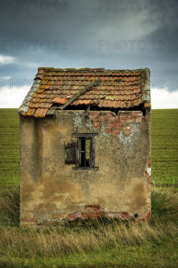 A small, dilapidated hut with a broken roof and cracked walls sits alone in a vast green field. Dark clouds loom overhead. Puy de Dome.Auvergne. France
