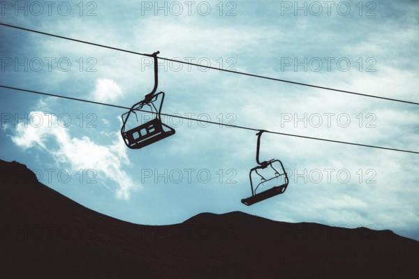 Two empty ski lifts hang from cables, silhouetted against a backdrop of a dark mountain and cloudy sky. The stillness captures the quiet essence of the ski resort at dusk. Auvergne Rhone Alpes. France
