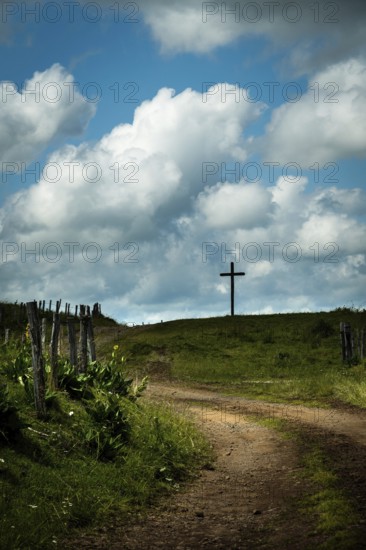 A winding dirt road leads the eye to a solitary cross. Cézallier, Auvergne Volcanoes Regional Natural Park, Puy-de-Dôme, Auvergne Rhone Alpes, France