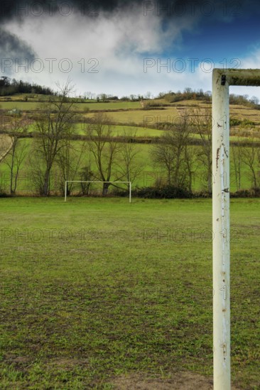 A football field is set against rolling hills and trees. The goalpost stands tall, framed by the vibrant greenery and dramatic clouds above, France