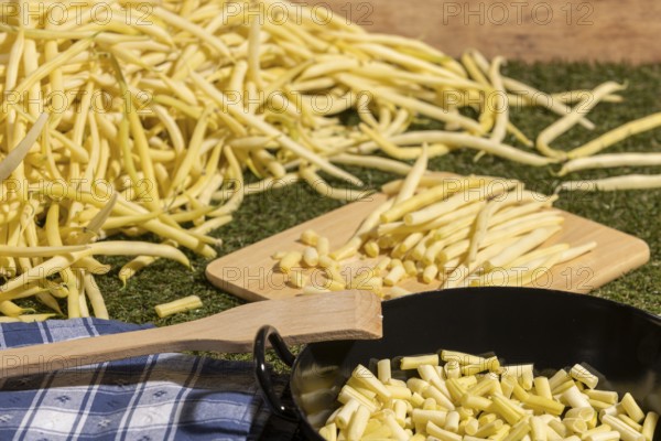 Yellow runner beans next to a cooking pot with sliced beans, with a wooden spoon on top