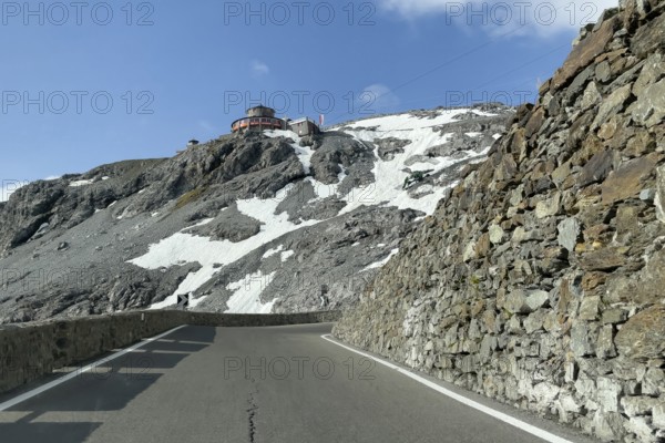 View in front of a narrow bend above the tree line in front of the Stelvio Alpine Pass road to the rocky mountain peak and the Hotel Tibethaus restaurant, Stelvio, Autonomous Province of Bolzano, South Tyrol, Italy