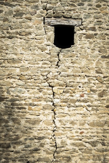 A weathered stone wall displays a prominent crack running vertically, featuring a small window. This structure, marked by time, showcases its rustic charm in a historical setting