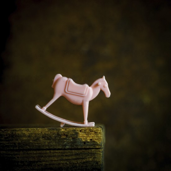 A small pink rocking horse toy sits on a weathered wooden shelf, gently illuminated in low light. The pastel color against the dark background evokes memories of childhood innocence and playfulness