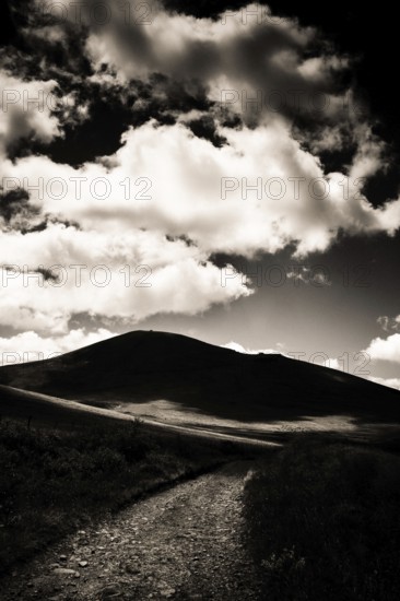 A winding path leads towards a dark mountain silhouette against a dramatic sky filled with billowing clouds. Sancy Massif, Puy de Dome, Auvergne, France