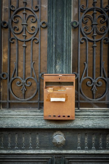 A rusty mailbox is affixed to a beautifully crafted wrought iron door. The intricate ironwork contrasts with the weathered wood. Auvergne. France