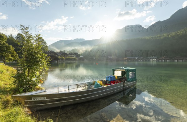 Beautiful morning at the Königssee in Schönau with a boat and boathouses in the background. Sunrise and beautiful wafts of mist over the Königssee in the basin