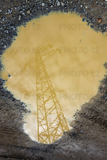 A power line tower is reflected in a large puddle of muddy water. The cloudy sky is visible, adding a moody atmosphere to the outdoor scene captured after rain