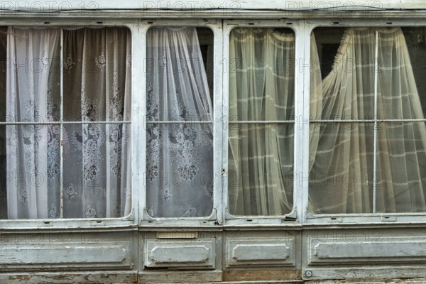 Light streams gently through patterned curtains hanging in a historic building, creating an atmosphere of calm and nostalgia. The weathered window frames hint at age and history. France