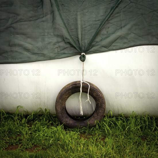 A weathered tire is tied to a green tarp, providing stability in a subtle, serene outdoor setting. Lush grass surrounds the scene, adding a touch of nature. France