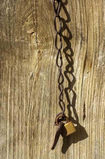 A close-up view of a wooden fence features a rusty chain and locked padlock. The sunlight enhances the texture of the weathered wood and metal, evoking a sense of tranquility. France