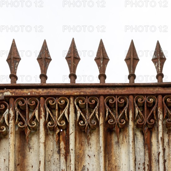 An old, weathered wrought iron fence features intricate scrollwork and pointed spikes atop its structure, showing signs of rust and age against a cloudy sky backdrop. France