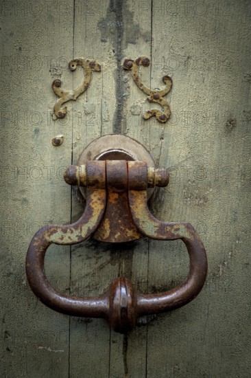 An old, rusty door knocker rests on a weathered wooden door, showcasing intricate designs and the effects of time. The worn surface adds to the unique character of the entrance