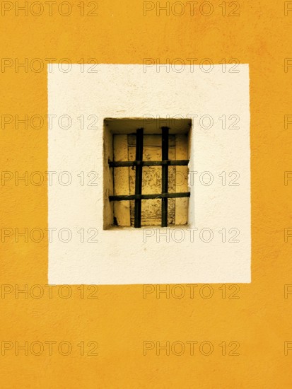 A small rectangular window with a black barred grille is centered in a white square, surrounded by a vibrant yellow wall. The contrast adds a lively touch to the urban architecture