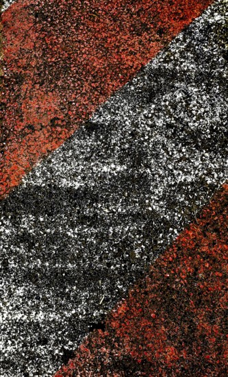 A close-up view of a textured pavement featuring bold diagonal stripes in red and black, showcasing worn surfaces and contrasting colors under midday sunlight