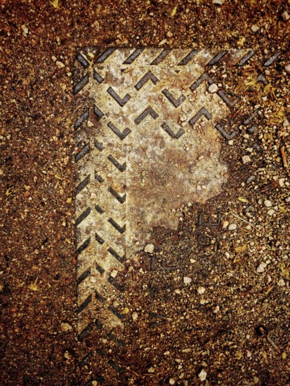 A worn metal cover is partially submerged in dirt and gravel, showcasing intricate patterns from erosion and weathering, highlighting nature's impact on human-made materials