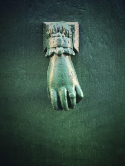 A unique green hand-shaped door knocker adorns a textured dark wall, adding a charming historical touch to the location. Its intricate design draws the eye and invites curiosity