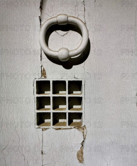A striking white door knocker adorns a weathered wall featuring small square openings arranged in an organized grid. The cracked surface adds to the vintage allure, exuding history and character