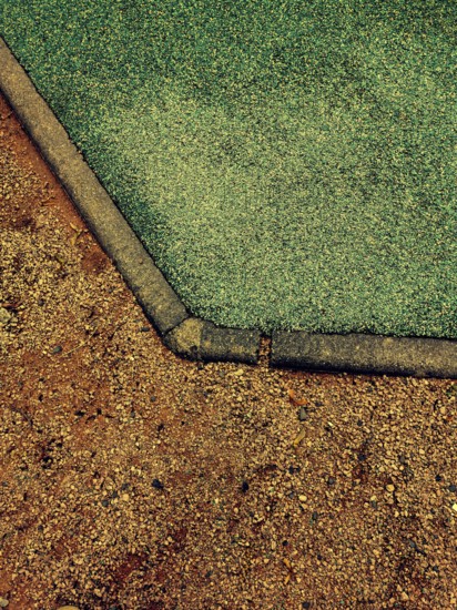 This detailed view captures the unique transition of a playground surface from vibrant green rubber to brown dirt during midday